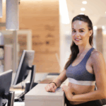 Gym member smiling at the front desk while staff uses gym management software on a computer