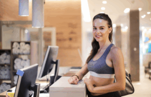 Gym member smiling at the front desk while staff uses gym management software on a computer