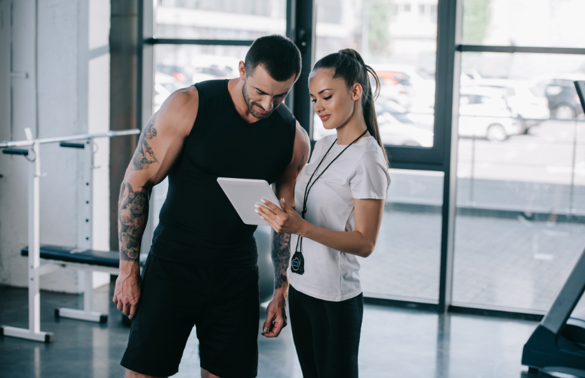 Personal trainer using fitness club management software on a tablet to review workout plans with a gym member.
